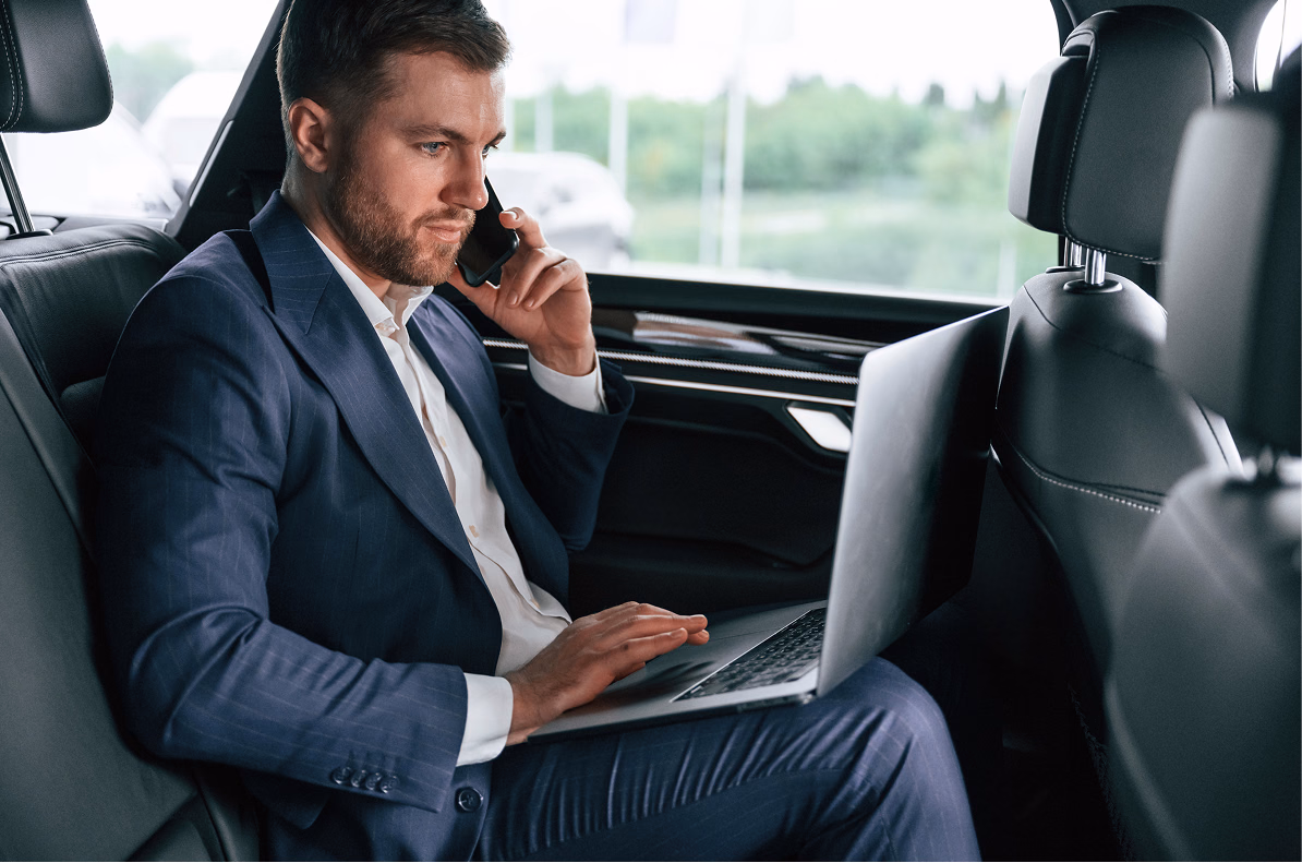 A businessman in a suit sitting in the back seat of a car while working on a laptop and talking on a smartphone.