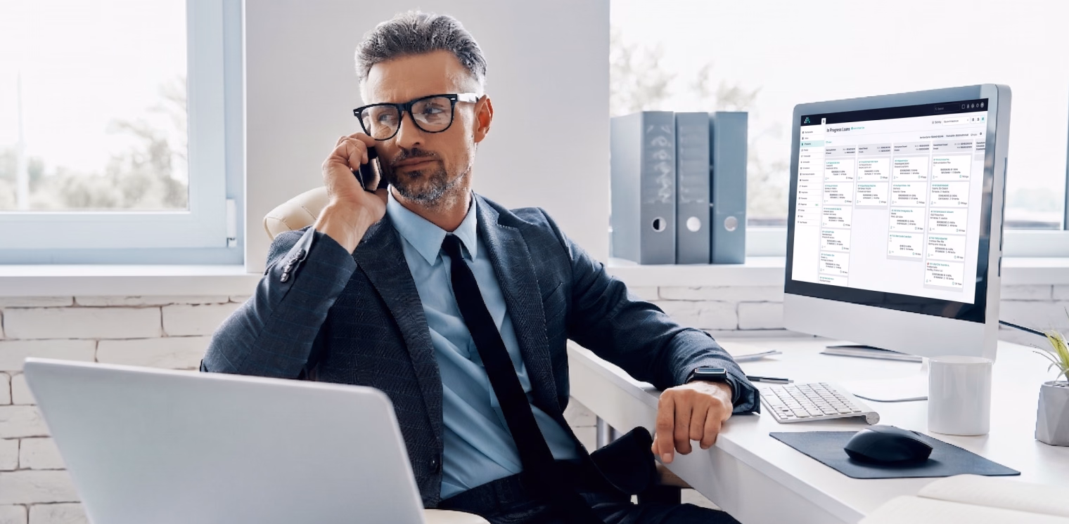 A professional man in a suit and glasses talks on his smartphone while sitting at his desk with a computer and laptop in a bright office.